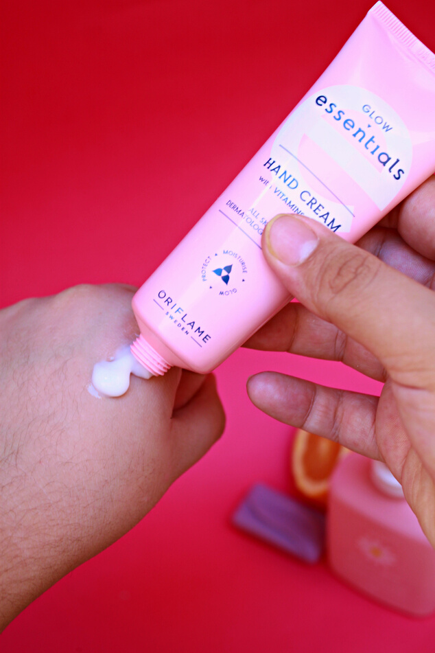 a UGC Creator hand holding a tube of the Glow Essentials Hand Cream with Vitamins E & B3 on a pink background